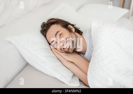 Jeune homme caucasien heureux souriant allongé dans un lit sous une couverture, atmosphère douillette Banque D'Images