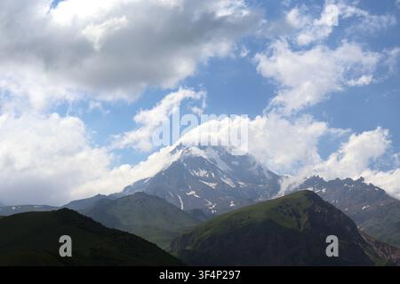 Sommet enneigé majestueux du mont Kazbegi partiellement enveloppé de nuages blancs duveteux avec des pentes montagneuses vertes luxuriantes, Géorgie Banque D'Images