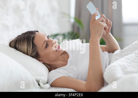 Jeune femme allongée dans un lit utilisant un smartphone, cadre domestique confortable, visage non identifiable Banque D'Images