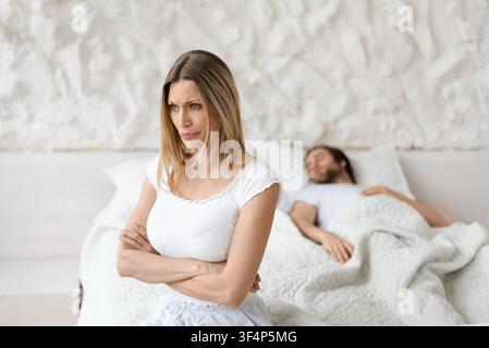 Femme blanche en détresse avec les bras croisés regardant au loin sur le lit, partenaire endormi, conflit relationnel Banque D'Images