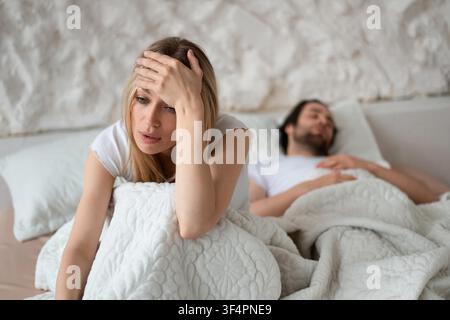 Femme blonde inquiète assise sur un lit à côté d'un homme détendu, se sentant stressée dans la chambre Banque D'Images