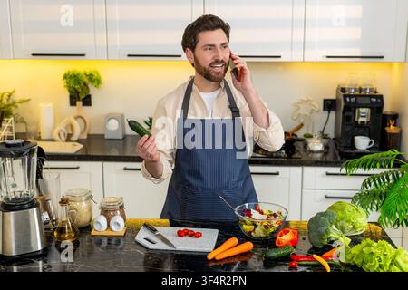 Homme d'âge moyen mélangeant salade de légumes et bouchée de dégustation tout en parlant sur smartphone dans la cuisine pour la santé. Jeune gars souriant pendant une alimentation saine appel de partage de succès et de motivation à la maison à l'intérieur. Banque D'Images