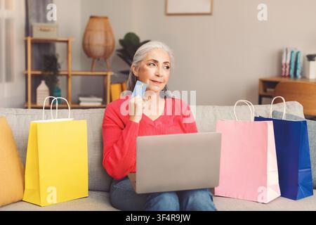 Femme caucasienne âgée réfléchie faisant ses achats en ligne avec un ordinateur portable, tenant une carte de crédit. Concept commercial Banque D'Images