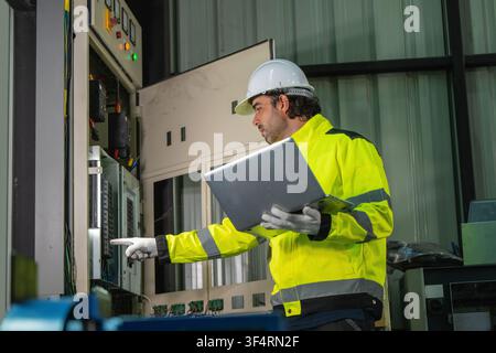 Ingénieur électricien masculin tenant un ordinateur portable inspectant un armoire électrique industrielle dans un entrepôt. Concept d'alimentation des machines lourdes, maintenance électrique Banque D'Images