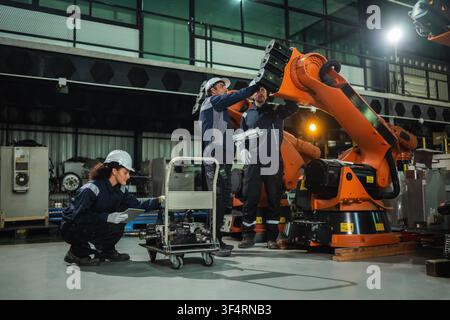 Équipe d'ingénieurs industriels discutant du contrôle d'une gamme de machines à bras robotiques orange tandis qu'une collègue vérifie des pièces détachées de servomoteur électrique. C Banque D'Images