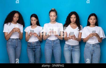 Cinq femmes du millénaire diverses utilisant des téléphones debout sur fond bleu Banque D'Images