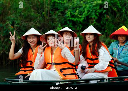 Quatre jeunes femmes souriantes portant des chapeaux coniques traditionnels et des gilets de sauvetage orange lors d'une excursion en bateau dans la région de Ninh Binh. Trang An, Vietnam. Banque D'Images
