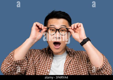 Homme asiatique surpris ajustant ses lunettes et souriant, fond de studio Banque D'Images
