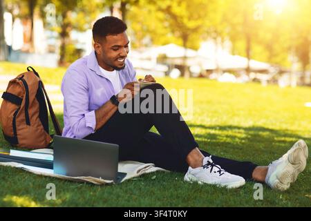 Homme noir souriant utilisant un smartphone assis sur l'herbe dans un parc, concept de détente Banque D'Images