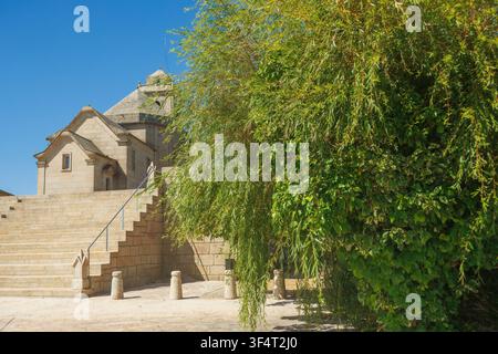 Vue horizontale de l'ancienne chapelle en pierre de San Fins avec de larges marches en granit et un feuillage de saule luxuriant sous un ciel bleu éclatant. Banque D'Images