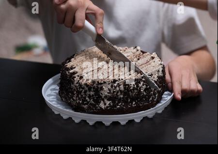 Un enfant coupe avec enthousiasme un gâteau au chocolat décadent, révélant ses couches tentantes tout en étant entouré de sa famille, illustrant la joie et la cohésion Banque D'Images