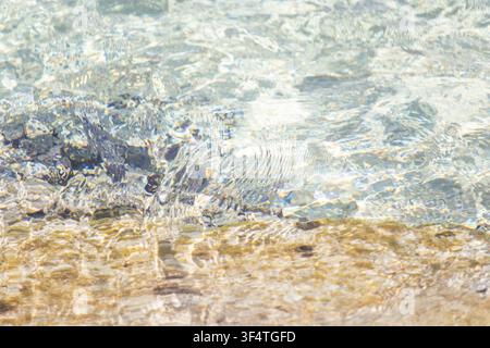 Eau de mer cristalline ondulant sur un fond sablonneux et rocheux par une journée ensoleillée, texture de surface de l'eau naturelle abstraite et réflexions de lumière Banque D'Images