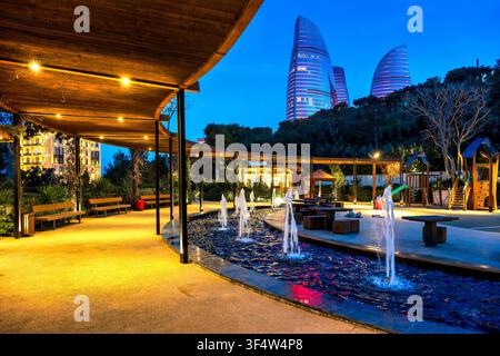 Fontaine illuminée et zone de pergola dans le parc de Chambarakand avec trois tours de flamme brillant à l'heure bleue au-dessus des cimes des arbres. Bakou, Azerbaïdjan. Banque D'Images