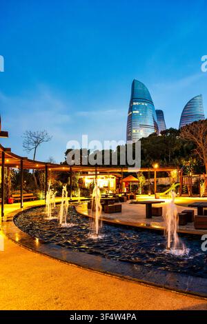 Fontaine illuminée et zone de pergola dans le parc de Chambarakand avec trois tours de flamme brillant à l'heure bleue au-dessus des cimes des arbres. Bakou, Azerbaïdjan. Banque D'Images