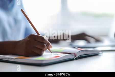 Une femme africaine méconnaissable prend des notes tout en travaillant sur un ordinateur portable au bureau Banque D'Images