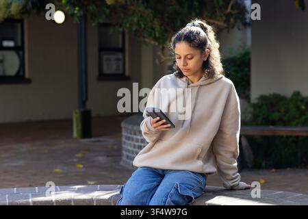 Adolescente afro-américaine assise sur un bac à fleurs en brique incurvé dans une cour pavée, tenant un smartphone Banque D'Images