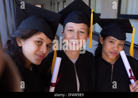 Trois étudiantes adolescentes posant dans un couloir avec des robes noires, des toges, des pompons jaunes, des diplômes Banque D'Images