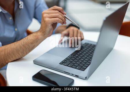 Grossissement des mains d'un homme d'affaires pointant sur un écran d'ordinateur portable avec un stylo argenté tout en travaillant à un bureau blanc. Smartphone au premier plan. Parfait f Banque D'Images