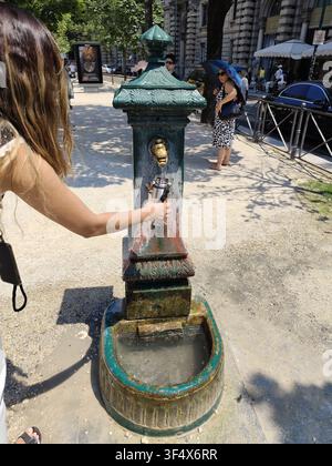 Femme remplissant une bouteille d'eau à une fontaine publique traditionnelle sur une rue italienne en été à Milan, Italie - 24 juin 2026 Banque D'Images