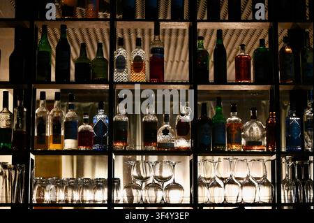 Une étagère de bar accueillante et atmosphérique présentant diverses bouteilles de liqueur et d'alcool vintage sur des étagères en bois éclairées, avec des verres à vin et à cocktail soigneusement disposés en dessous. Banque D'Images