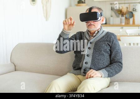 Un homme mature et enthousiaste portant des lunettes VR métaverse de réalité virtuelle à la maison. Grand-père touchant l'air pendant l'expérience VR sur un casque de réalité virtuelle. Simulation, jeu vidéo high-tech Banque D'Images