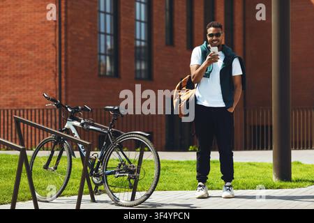 Jeune homme noir souriant utilisant un smartphone tout en se tenant à côté d'un vélo en plein air Banque D'Images
