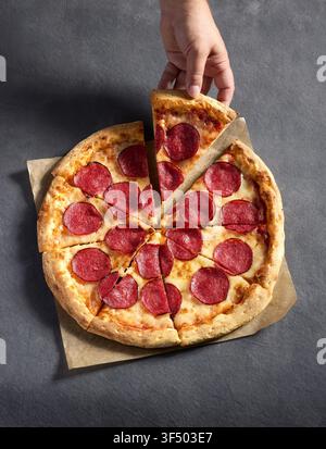 Gros plan d'une main tirant une délicieuse tranche d'une pizza entière au pepperoni et salami fraîchement cuite sur fond gris, tranches de salami épicées et un c Banque D'Images