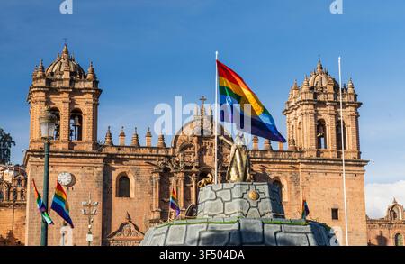Cusco, Pérou - 26 juin 2026 : Vue de la cathédrale de Cusco avec le drapeau arc-en-ciel de Cusco flottant devant une statue sous un ciel bleu clair. Banque D'Images