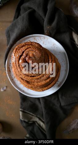 Une pâtisserie en spirale haute et épicée repose sur une assiette en céramique blanche, entourée d'un tissu texturé sombre et d'un éclairage latéral chaleureux mettant en valeur sa glace collante Banque D'Images