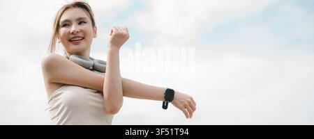 Belle femme asiatique étirant son bras dans une tenue de course fitness, femme souriante faisant de l'exercice sous un ciel lumineux tout en portant une montre connectée et Wi Banque D'Images
