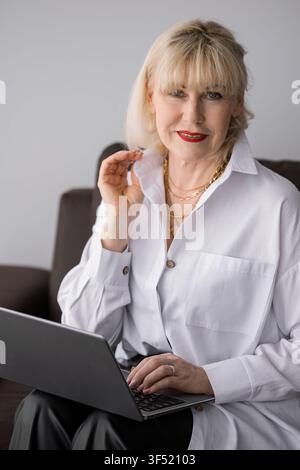 Femme senior élégante aux cheveux blonds et au rouge à lèvres rouge souriant avec assurance tout en tapant sur un ordinateur portable, représentant le travail à distance, l'apprentissage en ligne et Banque D'Images
