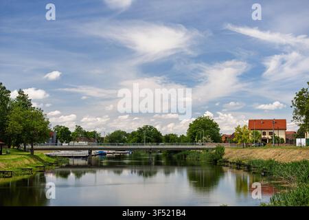 Scène de village paisible mettant en vedette une petite rivière avec des reflets, un pont en béton bas, des maisons aux toits rouges et des arbres verts luxuriants sous un ciel bleu avec clo Banque D'Images