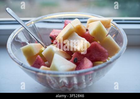 Un bol en verre transparent rempli de morceaux fraîchement coupés de pastèque rouge et de melon jaune, servi avec une petite cuillère en argent sur un rebord de fenêtre blanc. Banque D'Images