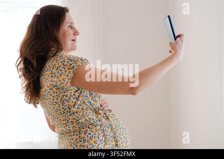 Une femme enceinte tient son smartphone pour un selfie ou un appel vidéo, partageant un moment heureux à l'intérieur. Une mère parle avec excitation et montre une joie joyeuse et enthousiaste ainsi qu'un bonheur maternel rayonnant à la maison Banque D'Images