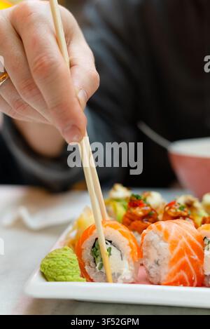 Grossissement d'une main d'homme prenant des sushis avec des baguettes. Des doigts habiles tenant un rouleau de saumon frais au-dessus d'une assiette appétissante. Photographie culinaire capturant le plaisir tactile de manger et la maîtrise des baguettes Banque D'Images