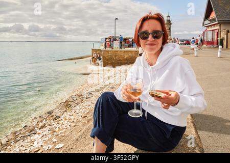 Femme en sweat à capuche blanc mangeant une huître fraîche avec un verre de vin sur la promenade côtière de Cancale, France, avec un fond marin. Banque D'Images