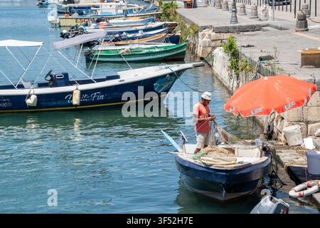 Syracuse, Italie - 8 juillet 2026 : Pêcheur réparant un filet sur un petit bateau de pêche dans un port par une journée d'été ensoleillée Banque D'Images
