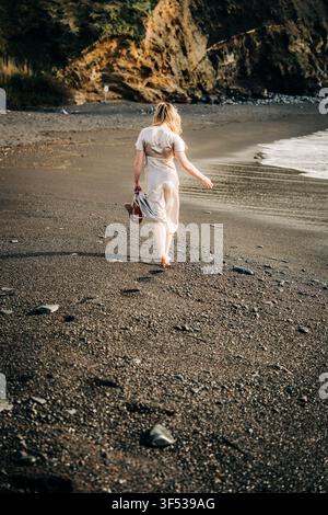 Femme blonde marchant pieds nus le long d'une plage de sable sombre Banque D'Images
