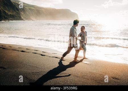 Couple heureux marchant le long d'une plage de sable noir au coucher du soleil Banque D'Images