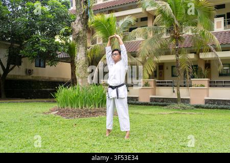 Jeune athlète karatéiste indonésien effectuant un étirement des bras au-dessus de la tête dans le cadre d'une routine d'échauffement avant un entraînement en plein air. Banque D'Images