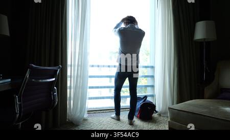 Jeune homme d'affaires fatigué en chemise debout près de la grande fenêtre dans la chambre d'hôtel moderne ou l'intérieur de l'appartement après un long voyage. Gestionnaire épuisé se reposant Banque D'Images