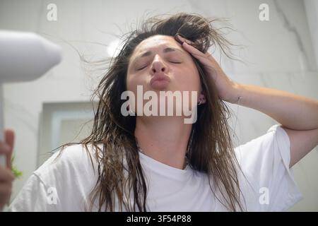 Femme profitant d'un moment de soins personnels tranquille à la maison, séchant ses cheveux après la douche avec un sèche-cheveux dans une salle de bain paisible. Banque D'Images