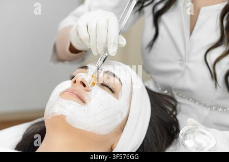 Close-up d'une procédure de soins de la peau où une cosmétologue applique un masque facial avec un pinceau sur le visage d'une femme dans un cadre de spa. La scène met en avant la détente, le bien-être et les traitements de beauté avancés Banque D'Images
