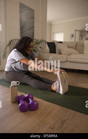 Femme afro-américaine adulte s'étirant sur un tapis vert à la maison avec des poids violets en vêtements de sport Banque D'Images