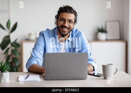 Joyeux Indien freelancer homme assis au bureau avec ordinateur portable, souriant à la caméra Banque D'Images