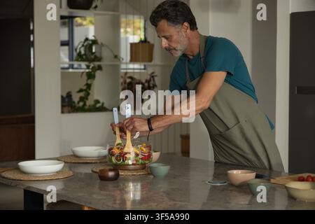 homme mûr en chemise bleu-vert et tablier vert olive lançant une salade sur l'îlot de cuisine avec un bol en verre transparent Banque D'Images