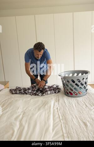 Homme adulte mûr se pliant dans la chambre, pliant une chemise à carreaux sur une couette blanche à côté d'un panier à linge gris Banque D'Images