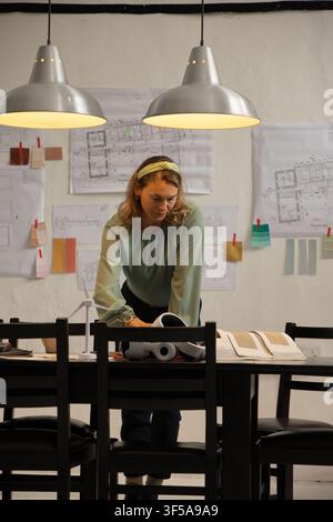 Architecte portant un chemisier vert, penchée sur une table sombre dans l'atelier en examinant des plans et un modèle Banque D'Images
