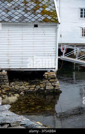 Ancien chantier naval en bois blanc construit sur des eaux calmes sur une fondation en pierre moussue sur la côte norvégienne, idéal pour un usage éditorial lié aux voyages et au patrimoine. Banque D'Images