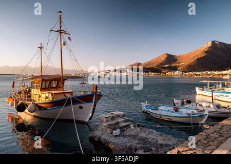 Elounda, Grèce – 2 septembre 2025 : Des bateaux en bois traditionnels et de petits navires de pêche sont amarrés dans le port au lever du soleil, avec la ville côtière et Banque D'Images